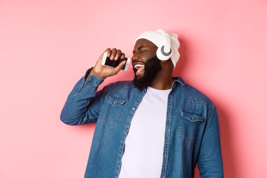 carefree african american man listening music headphones singing mobile phone as microphone standing pink background
